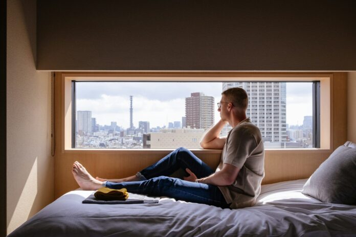 Photo by Johan Mouchet Man relaxes on a bed, gazing at a city view.